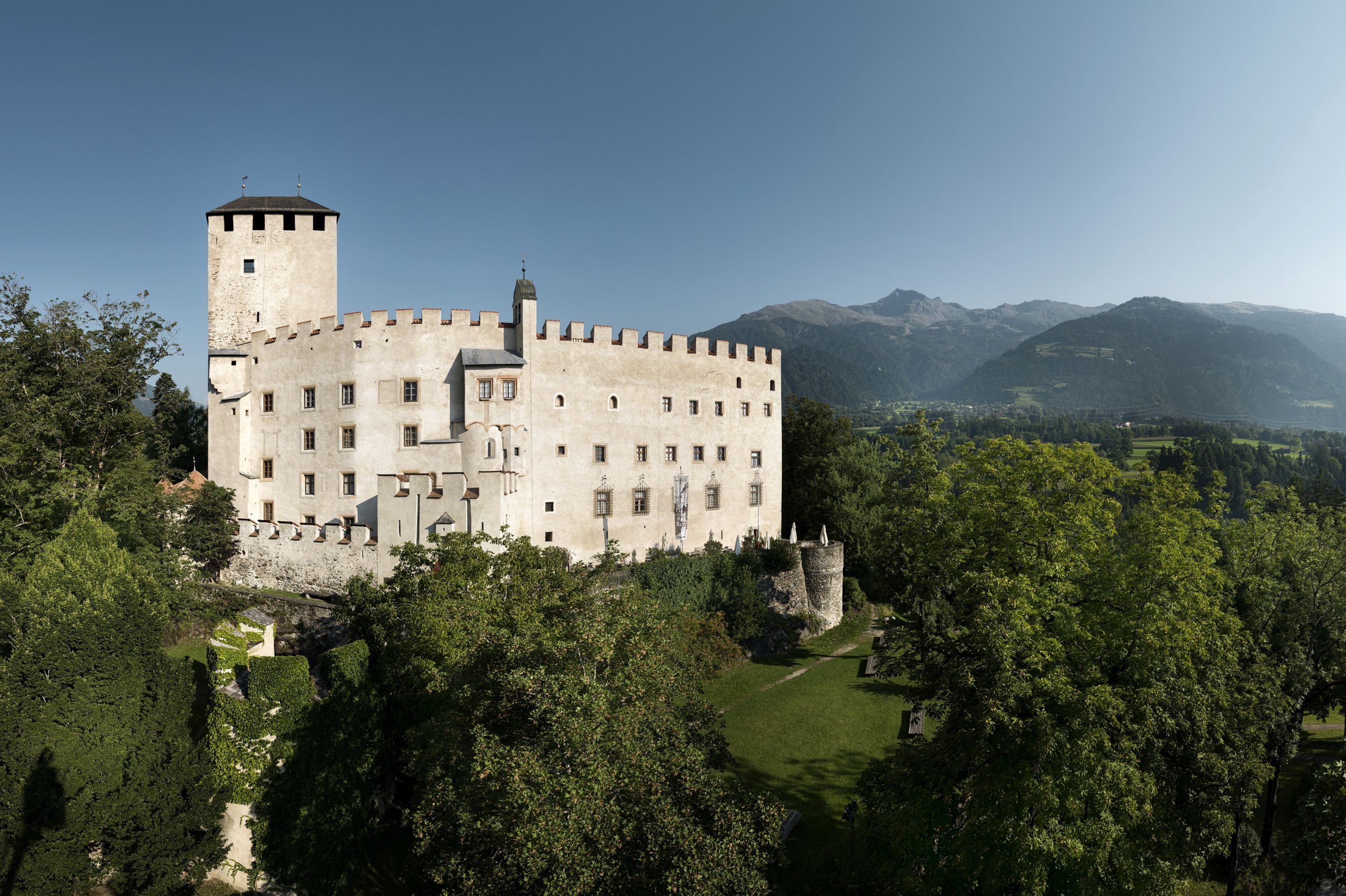 Schloss Bruck auf einer bewaldeten Erhebung, Bergpanorama im Hintergrund