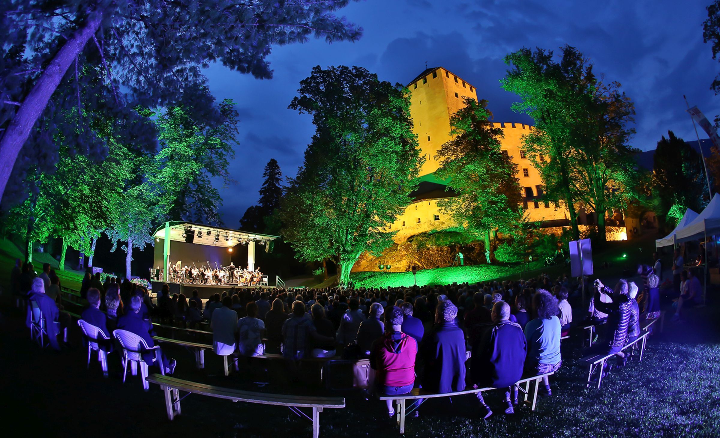 Konzertbühne am Abend mit bunten Lichtern und vielen Zuschauern, an der freien Luft vor dem beleuchteten Schloss Bruck